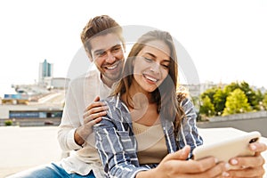 Image of young couple smiling while sitting outdoors with cellphone