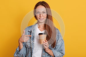 Image of young dark haired female pointing on paper coffee cup which holds in hand, woman dressed denim jacket and white t shirt,