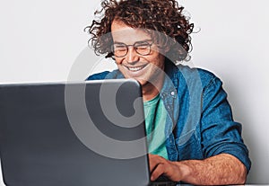 Image of young happy businessman with curly hair working on his laptop in office. Cheerful smiling student man sitting at his desk
