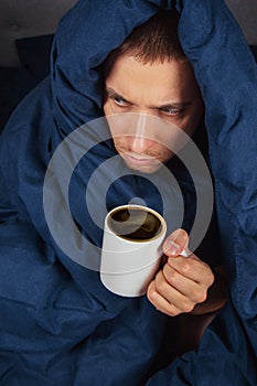 The image of a young man is sitting on a bed and covered with a dark blue blanket. He is holding a white cup of tea. The