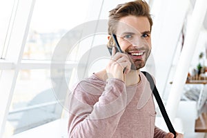 Image of young man talking on smartphone over bright window indoors