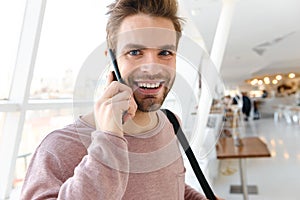 Image of young man talking on smartphone over bright window indoors