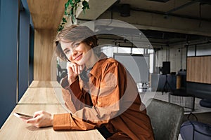 Image of young modern woman, employee in office, sitting at co-working space, working, using mobile phone, looking happy