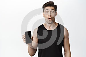 Image of young sportsman looking surprised, showing mobile phone screen, demonstrating app interface, gym website on