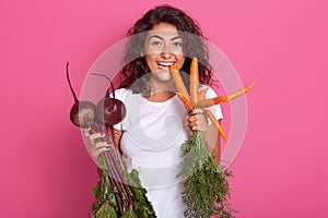 Image of young woman with dark wavy hair dressed white casual t shirt holding beets and carrots in hands, looking directly at