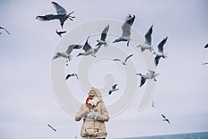 Image of young woman feeds seagulls on the sea. Pretty female wearing coat, scarf and watching flying seagulls by the sea in the