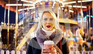 Image of young woman with glass of coffee in hands in park on background of carousel