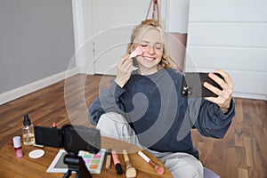 Image of young woman, vlogger taking selfie in her room, talking to her followers during online live stream, using