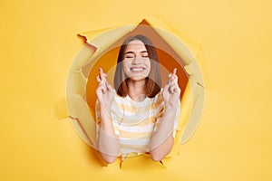 Imaged of smiling happy brunette woman stands in torn paper hole, standing with crosses fingers, posing with closed eyes, hope,