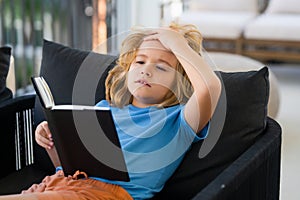 Imagination child. Cute kid in casual clothes reading a book while lying on a sofa in the room.