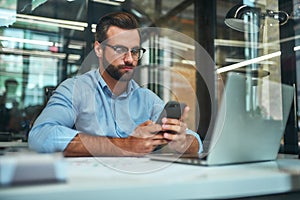 Important call. Portrait of young focused businessman in eyeglasses and formal wear looking at his smartphone while