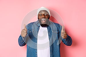 Impressed african-american hipster guy showing thumbs-up, approve and like offer, praising great choice, standing over