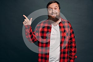 Impressed bearded hipster man in red checkered shirt, pointing left and smiling amused, sharing information where find great
