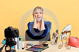 Impressed blogger sits in makeup room with palms near face, expresses happiness