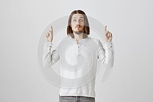 Impressed cute guy cannot believe his eyes. Portrait of handsome adult male model with beard and long hair pointing