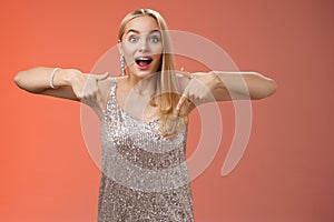 Impressed excited attractive glamour blond girl in silver glittering dress gasping thrilled pointing down glance camera
