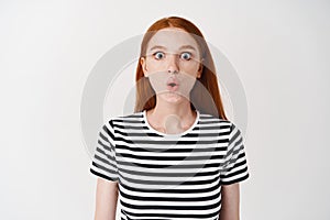 Impressed redhead girl stare at camera in awe, saying wow and looking astounded, standing over white background