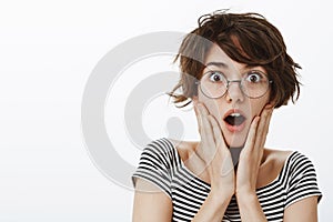 Impressed and shocked charming female with stylish short haircut in round trendy glasses, gasping and dropping jaw