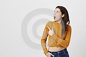 Impressed and surprised woman pointing at upper left corner while looking at it with widened eyes and opened mouth as if