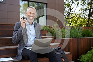 Improve your business. Joyful mature businessman smiling, making a call using his mobile phone while working on the