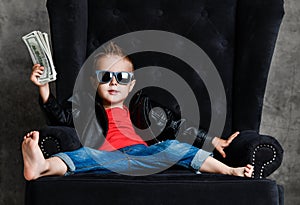 Impudent kid boy millionaire in leather jacket and red t-shirt is sitting lounging in armchair holding a bundle of cash