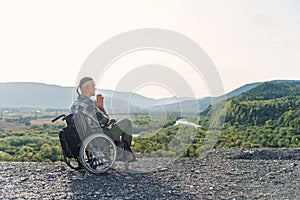 Likable incapacitated young guy sitting alone in wheelchair on the hill on beautiful nature background and praying. photo
