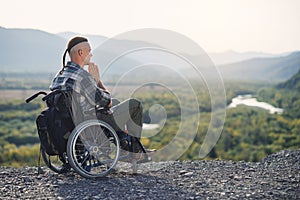 Likable incapacitated young guy sitting alone in wheelchair on the hill on beautiful nature background and praying. photo