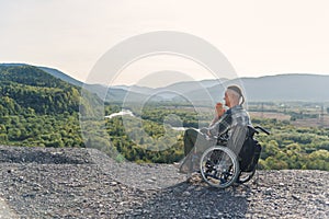 Likable incapacitated young guy sitting alone in wheelchair on the hill on beautiful nature background and praying. photo
