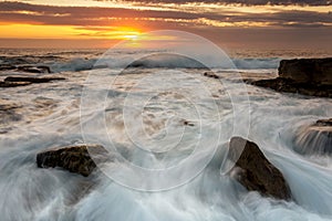 Incoming waves surge over rocks with a golden orange sunrise