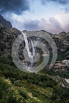 An incredible view of a majestic Shdugra waterfall, high in the Caucasus Mountains, rocky slopes, a rushing stream. Green herbs