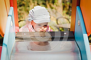 Indecision blond hair toddler girl in funny hat on a playground on a sliding in a public park