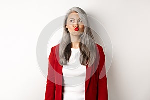 Indecisive asian businesswoman pucker lips and looking thoughtful at upper left corner, standing over white background