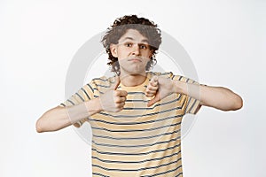 Indecisive young man showing thumbs up and thumbs down, looking confused about his decision, standing over white