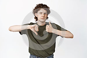 Indecisive young man shows thumbs up and thumbs down, like or dislike, looking doubtful at camera, standing over white