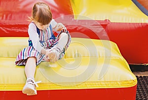 An independent Little tired girl is sitting on a children`s inflatable trampoline in an amusement park. A small child puts on his