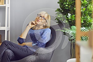 Independent mature woman sitting in armchair at home, thinking and looking at camera
