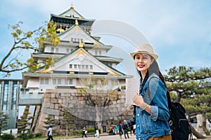 Independent travel in japan lifestyle concept. woman traveler holding guidebook visiting osaka castle alone carrying backpack and photo