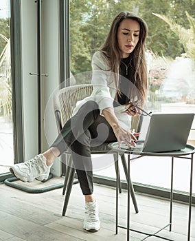 Independent women portrait. Young businesswoman working on a silver laptop, sitting near big window in an office