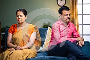 Indian angry middle aged couple sitting on sofa in opposite direction at home - concept of conflict, family problems and