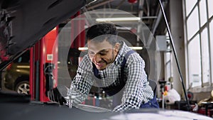 Indian auto mechanic examining broken car engine, standing near his car with open hood in a car repair shop