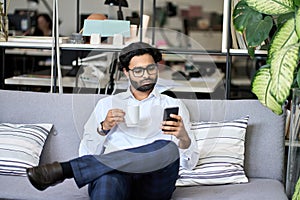 Indian business man using mobile phone reading news drinking coffee in office.