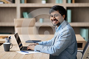 Indian businessman using laptop sits at desk smiling to camera