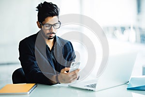 Young Indian CEO of company in his business office at desk, reading text on smartphone