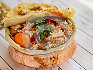 Indian Chicken Biryani with tomato, coriander, cashew nut and bread dum served in copper pot dish isolated on wooden table top