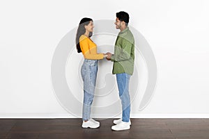 Indian couple holding hands and smiling to each other against white wall