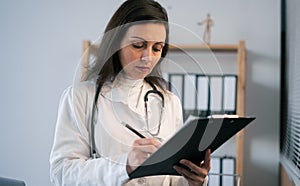 Indian doctor in white coat holding clipboard and writing diagnosis, close up. Quality medical services. Medicine and