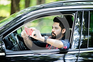 Indian driver taking photo with camera smartphone driving in car. Happy man taking picture with smart phone camera out window of