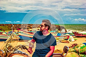 An Indian ethnicity man with sunglasses, stubble and black hair sitting on a cruiser bike against seashore and cloudy blue sky.