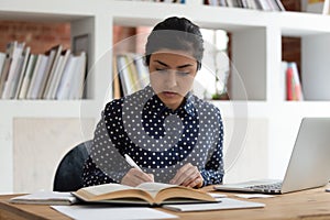 Indian ethnicity student woman writing notes reading textbook studying indoors
