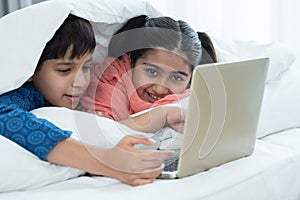 Indian family, brother and sister with traditional clothes smiling using laptop in bedroom at home, two children lying on bed